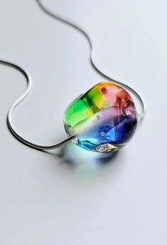 "This pendant features a rainbow hued handmade flamework glass pebble strung on a sterling silver snake chain. Each tactile 'pebble' is completely unique, built from layers of transparent colours encased in clear glass and handshaped in the torch flame. All glass is kiln annealed for durability before combining with an 18\" or 20\" sterling silver snake chain." Silver Murano Glass Round Pendant Necklace, Unique Iridescent Glass Necklaces, Modern Handmade Rainbow Jewelry, Handmade Rainbow Glass Jewelry, Rainbow Glass Jewelry Gift, Rainbow Glass Jewelry For Gifts, Modern Rainbow Colored Jewelry For Gifts, Modern Rainbow Jewelry Gift, Modern Rainbow Jewelry For Gifts