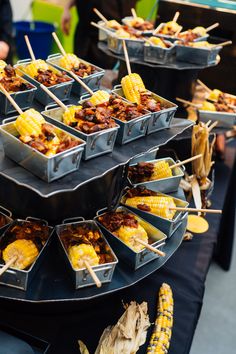 several trays of food are arranged on a table with corn and skewers