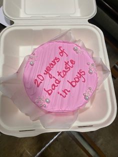 a pink frosted birthday cake in a plastic container with writing on the side and decorations around it