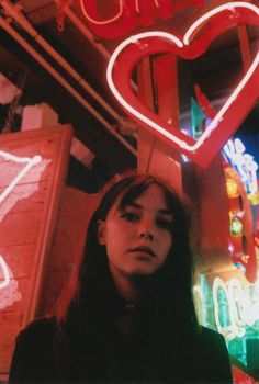 a woman standing in front of a neon sign with a heart hanging from it's side