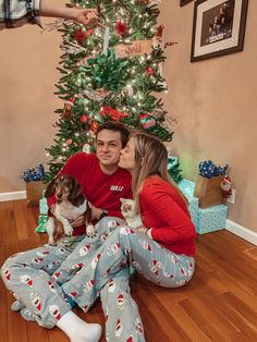 a man and woman sitting on the floor kissing in front of a christmas tree with their dog