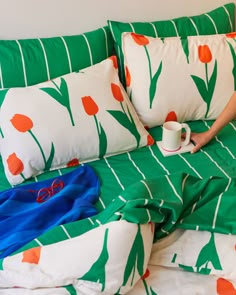 a person sitting on a bed with green and white sheets, holding a coffee cup