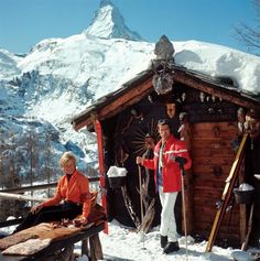 two people standing in front of a small cabin with skis on the ground and snow covered mountains behind them