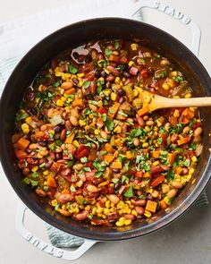 a skillet filled with beans and vegetables on top of a white table cloth next to a wooden spoon