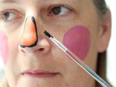 a woman with her face painted orange and pink, holding a paintbrush in front of her eye