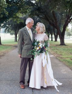 an older man and woman standing next to each other on a road with trees in the background