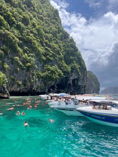 many people are swimming in the ocean with boats and cliffs on the shore behind them