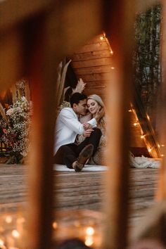 a man and woman sitting on the ground in front of a wooden structure with lights