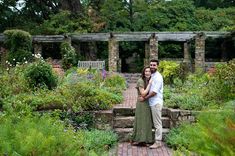 a man and woman standing next to each other on a brick walkway surrounded by greenery