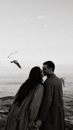 a man and woman standing next to each other near the ocean with seagulls flying overhead