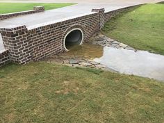 a brick wall with a drainage pipe running into the ground next to grass and water