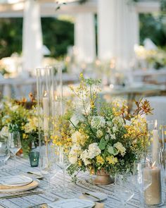 the table is set with white and yellow flowers in vases, candles, and napkins