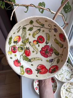 a hand holding a bowl with tomatoes and peppers painted on the side, in front of plates