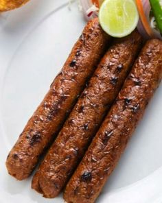 two sausages on a white plate next to some vegetables and a pickle slice