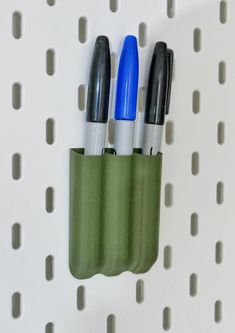 three pens in a green holder on a white pegboard with perforated holes