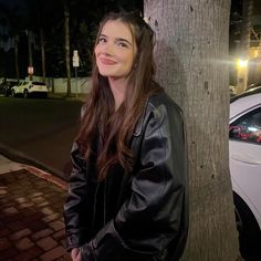 a woman standing next to a tree in front of a white car on the street