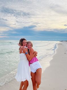 two women hugging on the beach with their arms around each other and one woman is wearing a white dress