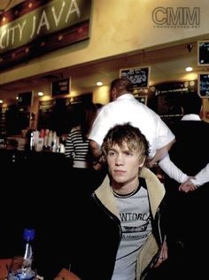 a young man sitting at a table in front of a bar with people behind him