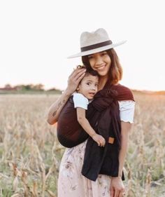 a woman in a hat holding a baby wearing a brown and white carrier while standing in a field