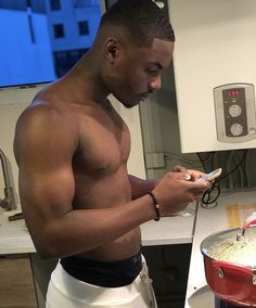 a shirtless man using his cell phone while cooking in a pot on the stove