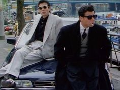 two men in suits and sunglasses sitting on the hood of a car near a marina