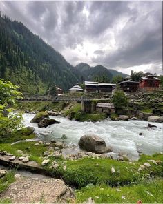 a river running through a lush green forest filled with lots of rocks and grass under a cloudy sky