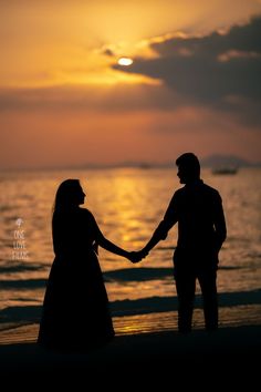 a couple holding hands while standing on the beach at sunset with the sun setting in the background