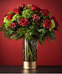 a vase filled with red roses and greenery on top of a wooden table next to a red wall