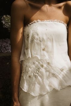 a woman wearing a white dress with flowers on it's chest and back, standing in front of a bush