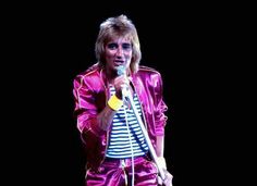 a man in pink is holding a microphone and wearing purple pants with stripes on them