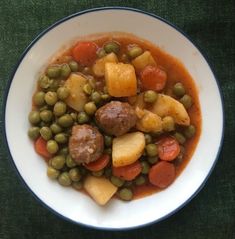 a white bowl filled with peas, potatoes and meatballs on top of a green table