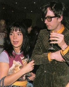 a man and woman standing next to each other holding wine glasses in their hands while one holds a cat