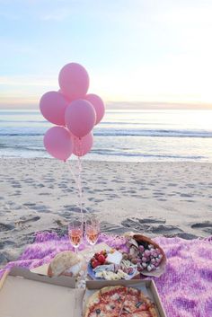 a pizza and some pink balloons on a blanket at the beach with food in front of it