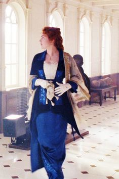 a woman in blue dress and white gloves walking through a hall with other people behind her