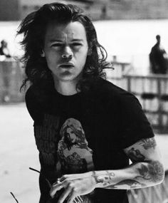 a man with long hair and tattoos on his arm holding a skateboard in front of him