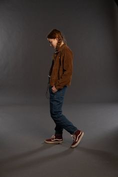 a man standing in front of a gray background wearing a brown jacket and blue jeans