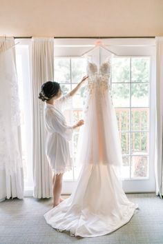 a woman standing in front of a window next to a wedding dress