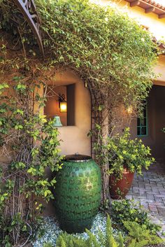 a large green vase sitting in the middle of a garden next to a building with ivy growing around it