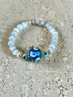 Evil Eye Bracelet featuring a Venetian Glass focal bead measuring approximately 20MM in shades of blue, white and yellow, accented with 6MM evil eye beads, gold plated bead caps and 8MM smooth round cat's eye beads.  Bracelet is strung on stretch cord and measures approximately 7 1/2 inches.  Handmade. GIFT BOXED White Glass Beaded Bracelets With Round Beads, Handmade White Glass Beaded Bracelets, White Glass Beaded Bracelets, White Beaded Bohemian Evil Eye Bracelet, Bohemian White Evil Eye Bracelet With Colorful Beads, White Bohemian Beaded Evil Eye Bracelet, White Round Bohemian Stretch Bracelet, White Bohemian Stretch Bracelet, White Evil Eye Jewelry For The Beach