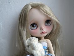 a doll with blonde hair holding a white teddy bear in her lap and looking at the camera