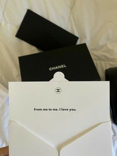 a person holding up a chanel card on top of a white bed with black pillows