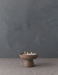 three lit candles sitting on top of a wooden bowl in front of a gray wall