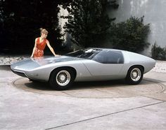 a woman in an orange dress standing next to a silver sports car on a driveway