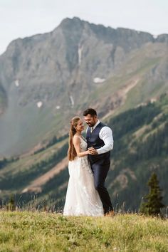 a bride and groom standing on top of a grass covered hill with mountains in the background