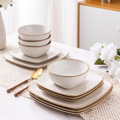 white and gold dinnerware set on a table with flowers in the vase behind it