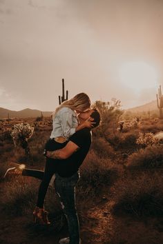 a man holding a woman in his arms while standing in the desert with cacti behind him