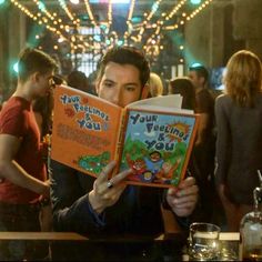 a man sitting at a bar reading a book with other people in the back ground