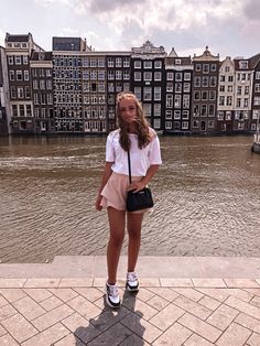 a woman standing on the side of a river with buildings in the back ground and water behind her