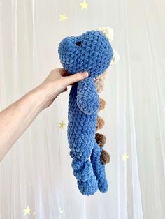 a hand holding a blue crocheted stuffed animal in front of a white curtain