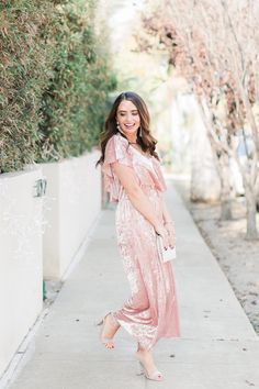 a woman standing on the sidewalk wearing a pink velvet dress and heels with her hands in her pockets